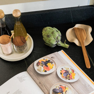 Mai Lidded Cellar & Spoon on Marble Lazy Susan next to Cruets, Cookbook, Spoon Rest, Artichoke, Linen Tea Towel, and Garlic Clove